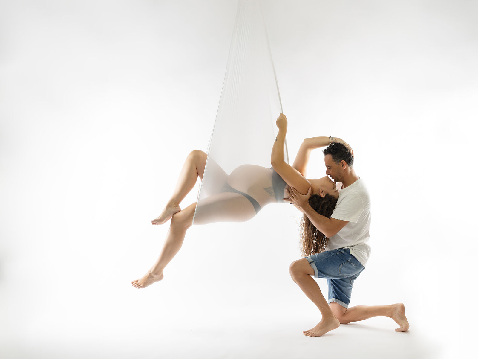 Mujer embarazada columpiada en una gasa transparente mientras su pareja la abraza de rodillas, en una sesión artística con fondo blanco realizada en estudio profesional en Cartagena por Torres & Torres Fotografía