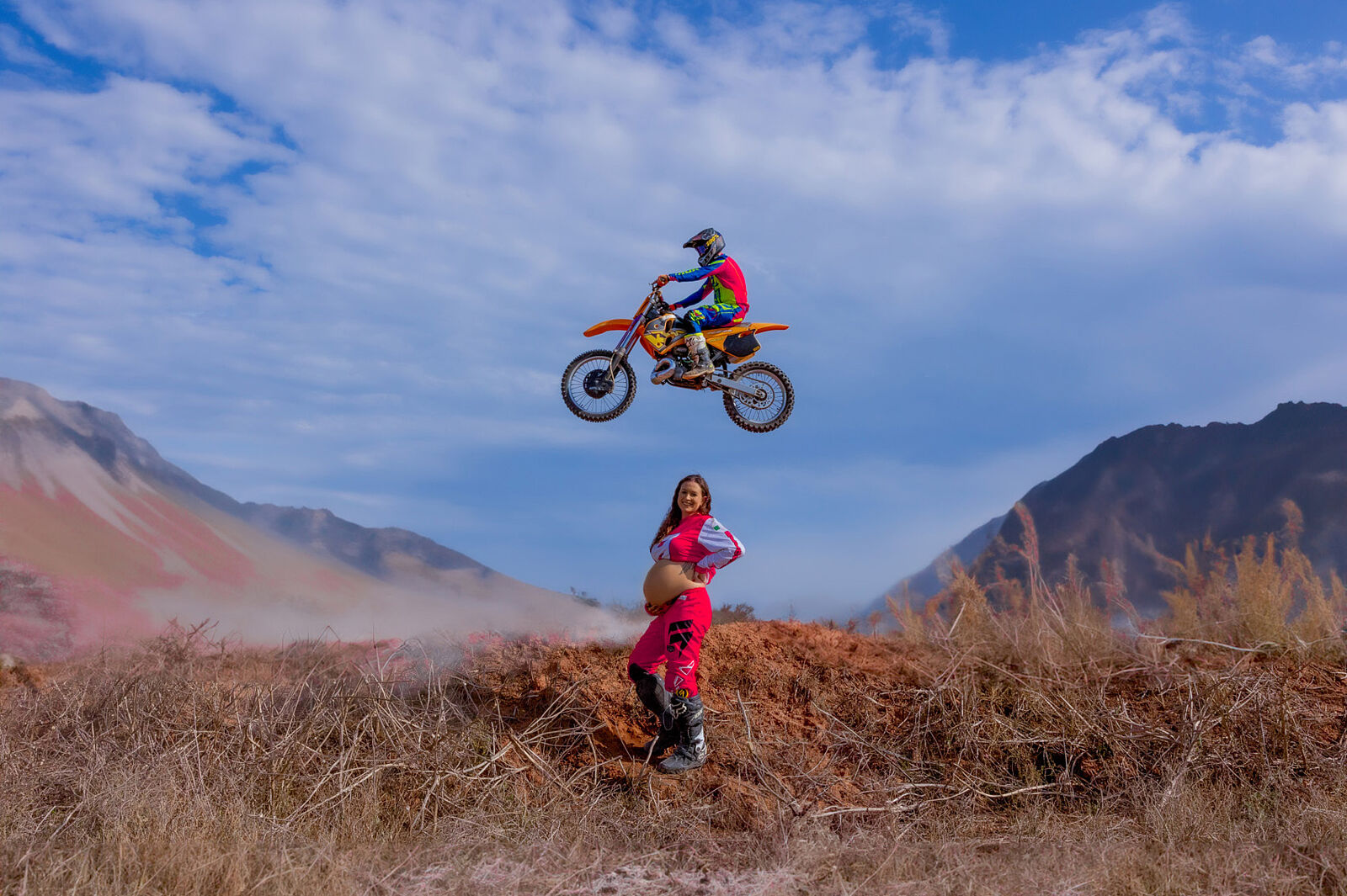 Mujer embarazada con mono de motocross posa sobre terreno montañoso mientras una moto vuela sobre ella en salto acrobático, durante sesión creativa en exteriores cerca de Cartagena por Torres & Torres
