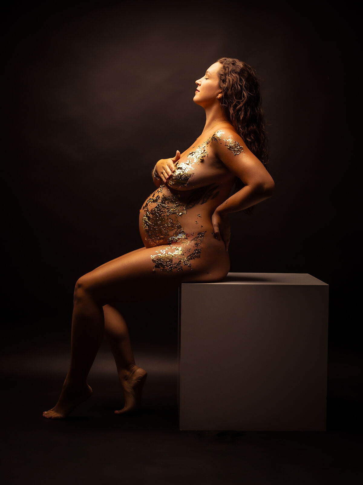 Mujer embarazada sentada en pose elegante, con el cuerpo decorado con pan de oro y luz cálida sobre fondo oscuro, durante una sesión artística en estudio en Cartagena realizada por Torres & Torres Fotografía