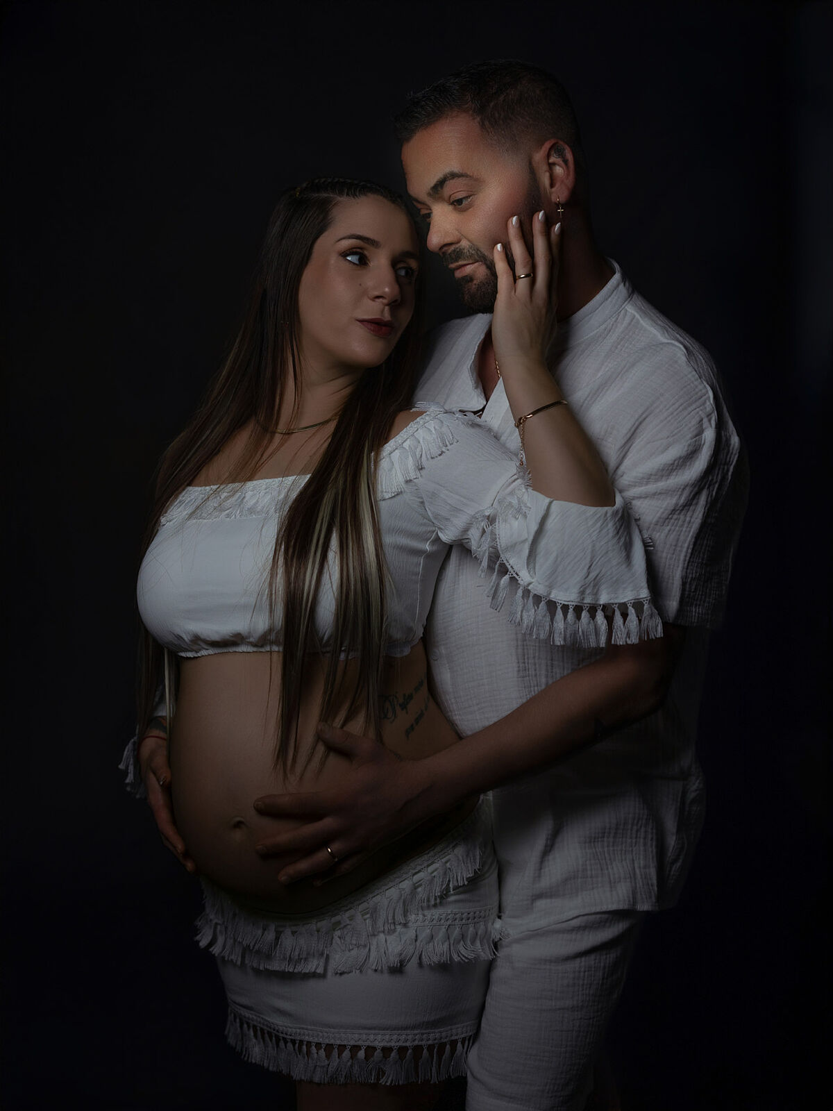 Retrato de pareja durante sesión de embarazo en estudio, ambos vestidos de blanco, con iluminación suave que resalta la conexión emocional, realizado en Cartagena por Torres & Torres Fotografía          Preguntar a ChatGPT