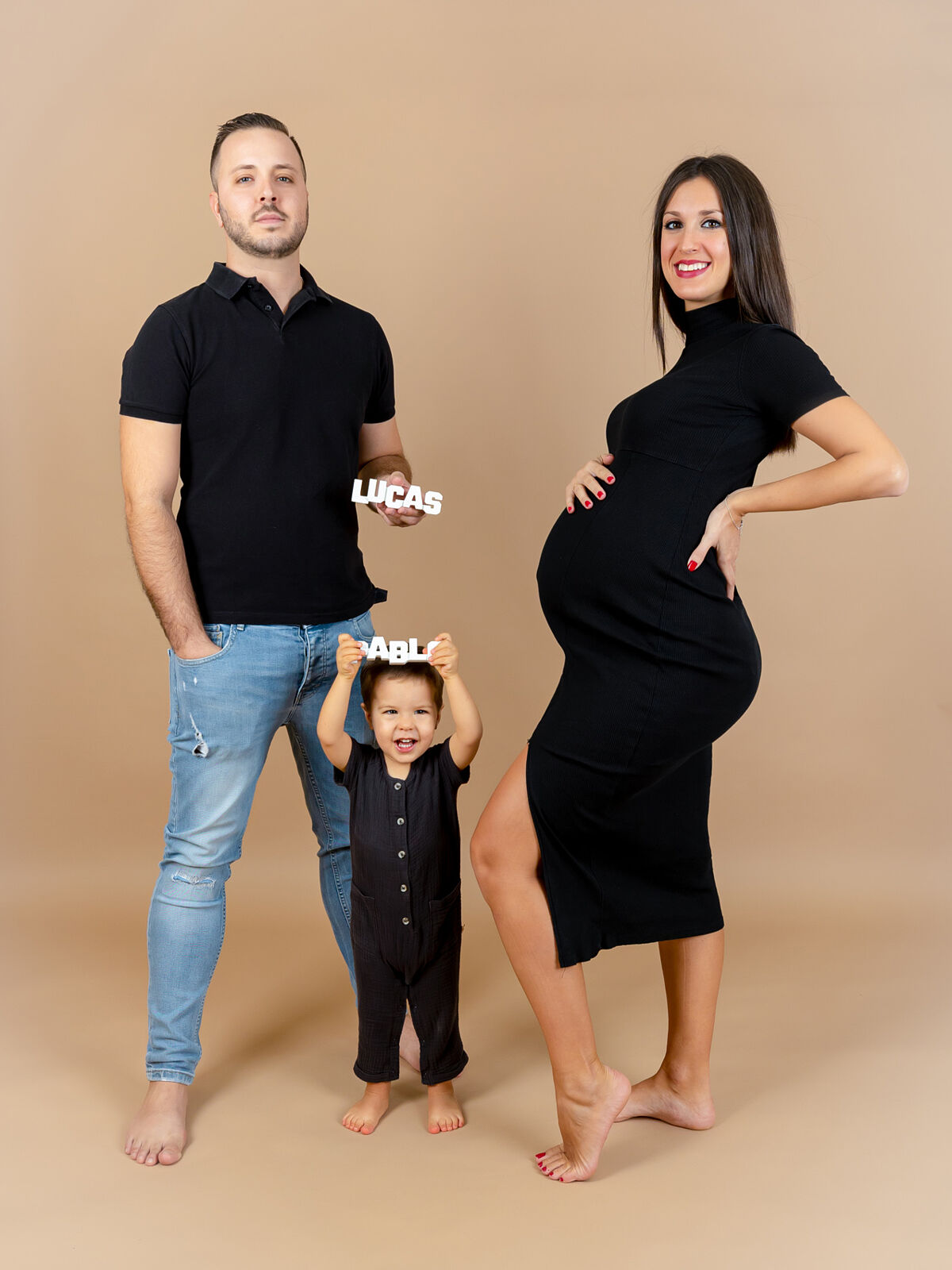 Mujer embarazada junto a su pareja e hijo pequeño, todos vestidos en tonos oscuros sobre fondo beige, en una sesión familiar alegre realizada en estudio profesional en Cartagena por Torres & Torres Fotografía          Preguntar a ChatGPT