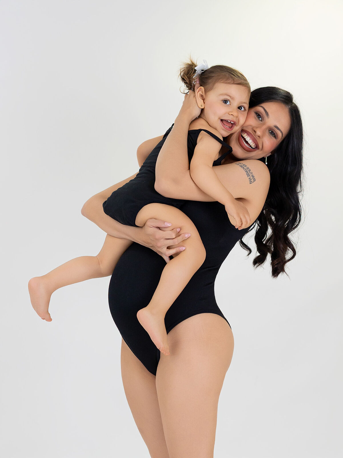 Madre embarazada vestida de negro sostiene con ternura y complicidad a su hija pequeña, ambas sonrientes en un estudio de fondo blanco en Cartagena, capturadas por Torres & Torres Fotografía