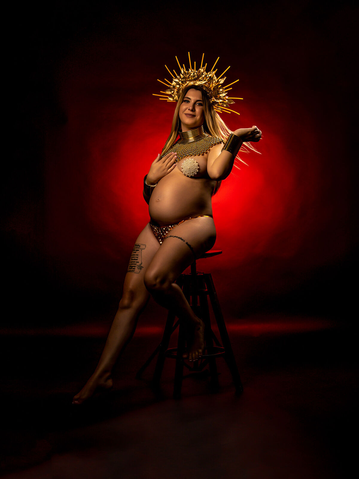 Mujer embarazada con corona dorada y estilismo inspirado en figuras divinas, sentada sobre taburete con fondo rojo dramático, en sesión artística de estudio en Cartagena por Torres & Torres Fotografía