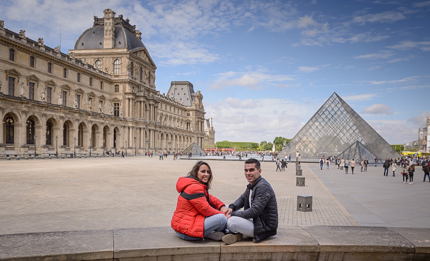 Casal de namorados sentados em cima do muro e de fundo tem o Museu do Louvre