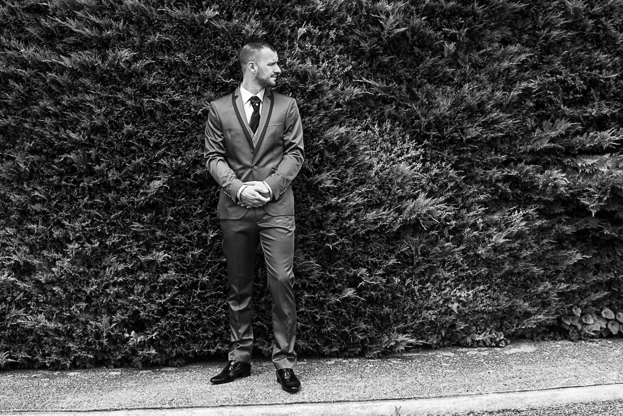 O noivo Kévin a olhar para o lado e de fundo arbustos