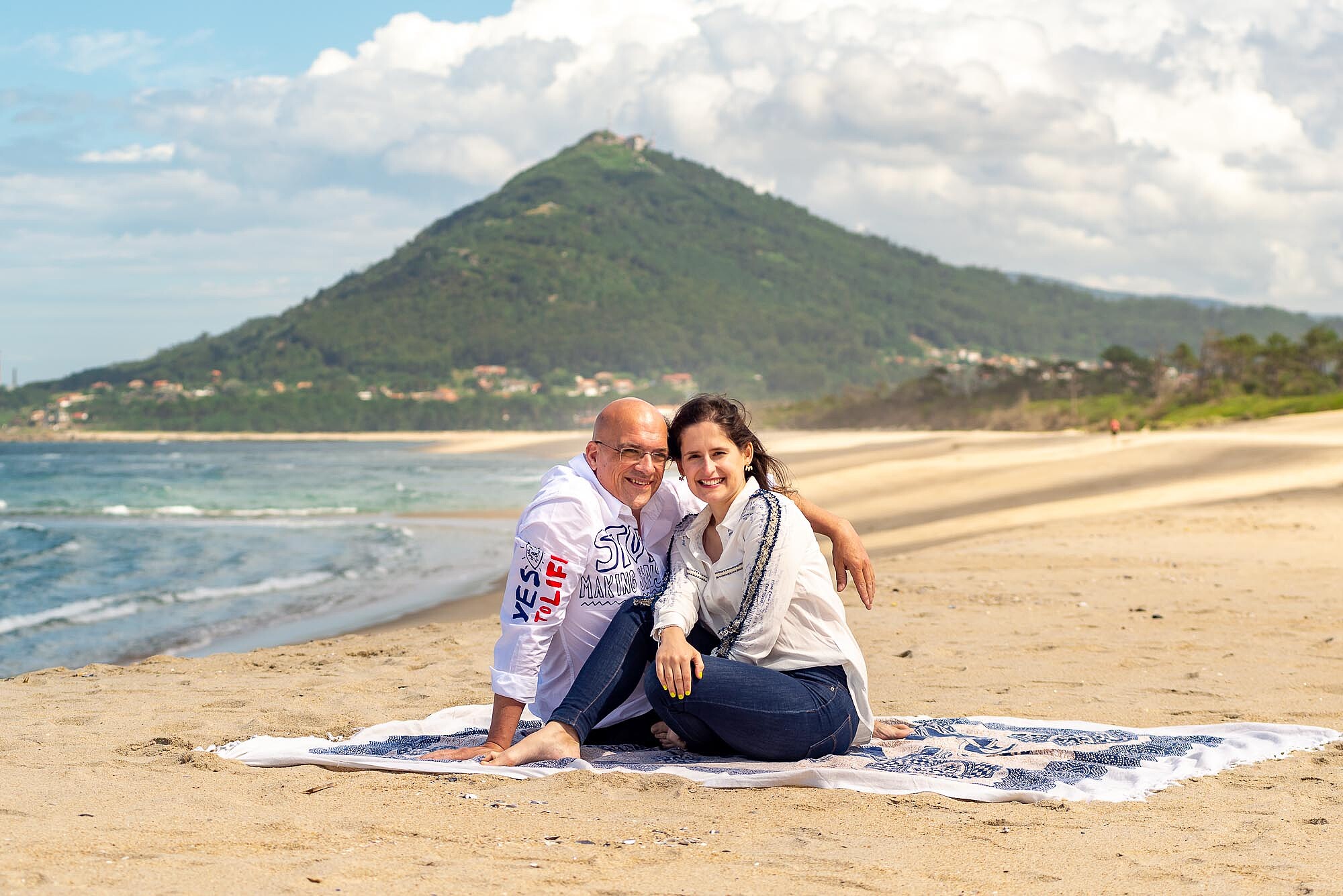 Noivos com a cabeça encostada uma na outra sentados na toalha de praia a beira mar