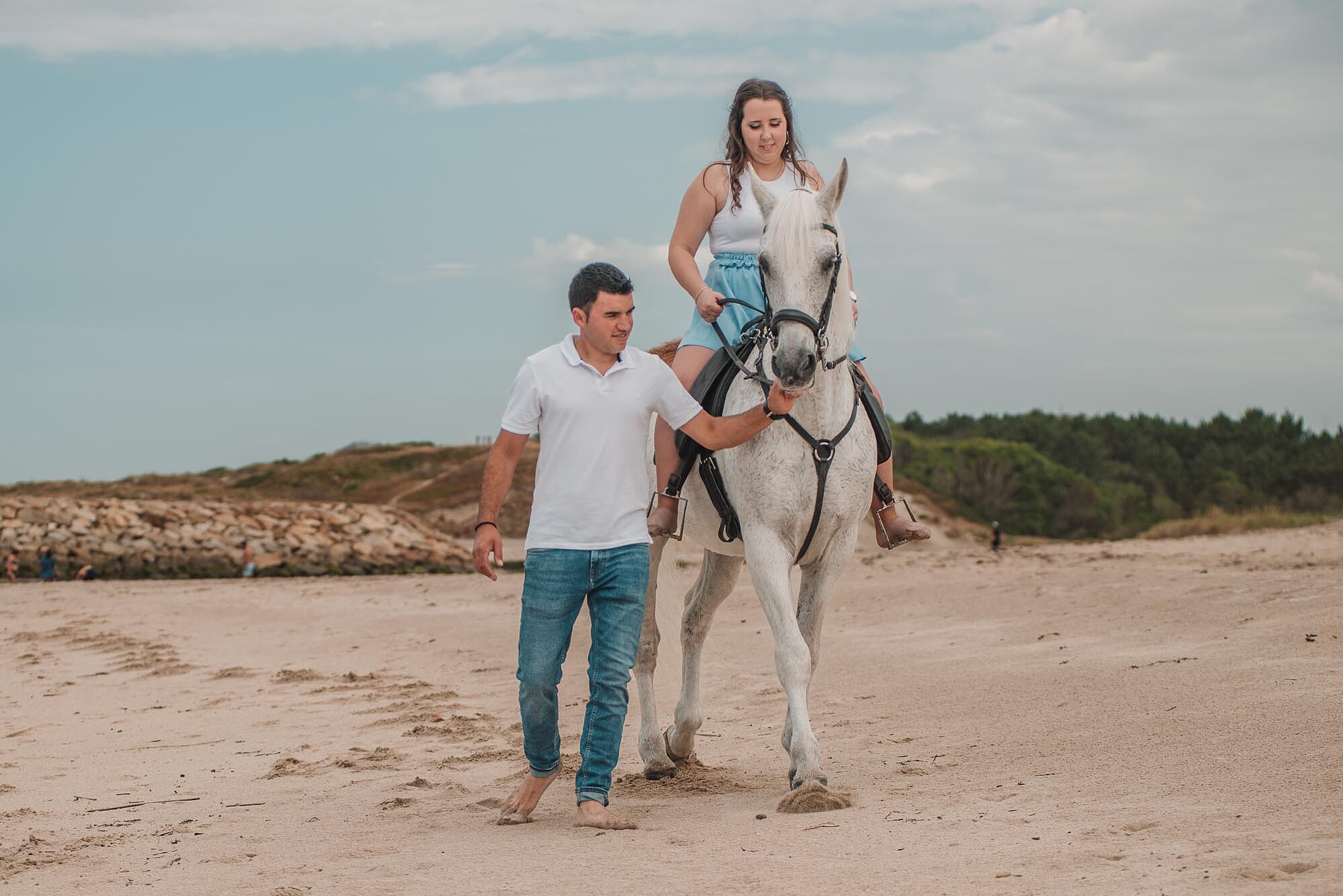 Mulher em cima do cavalo a caminhar na areia enquanto o namorado est&aacute; ao lado
