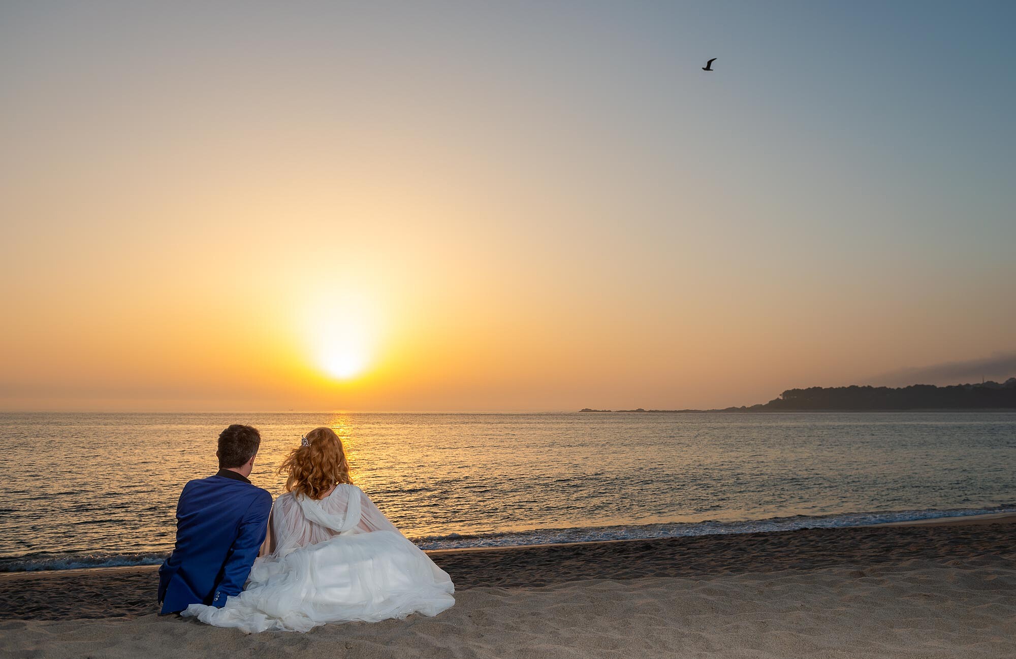 Casal sentado na areia a olhar para o por do sol