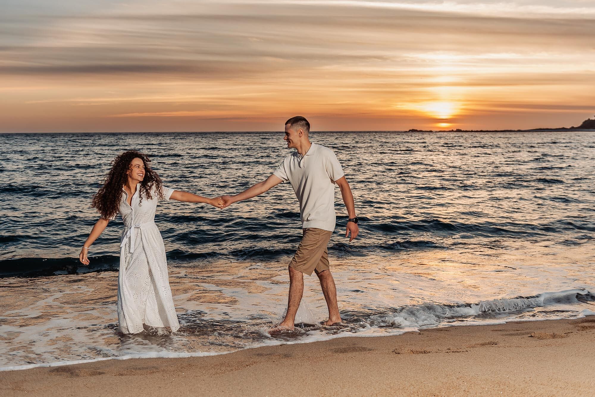 casal a brincar de maos dadas na beira da agua ao pôr do sol