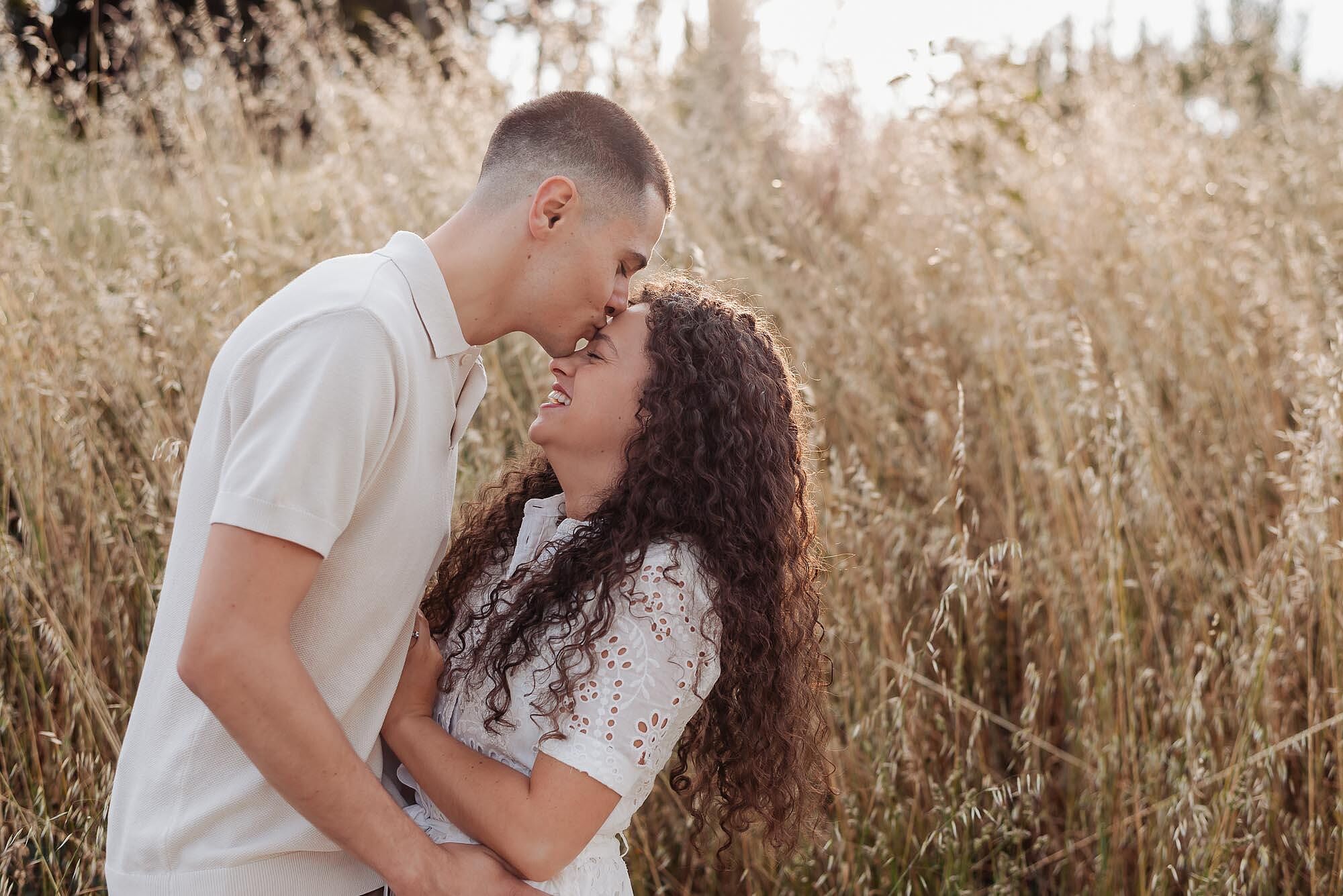 homem beija a testa da mulher enquanto ela ri num campo de ervas douradas