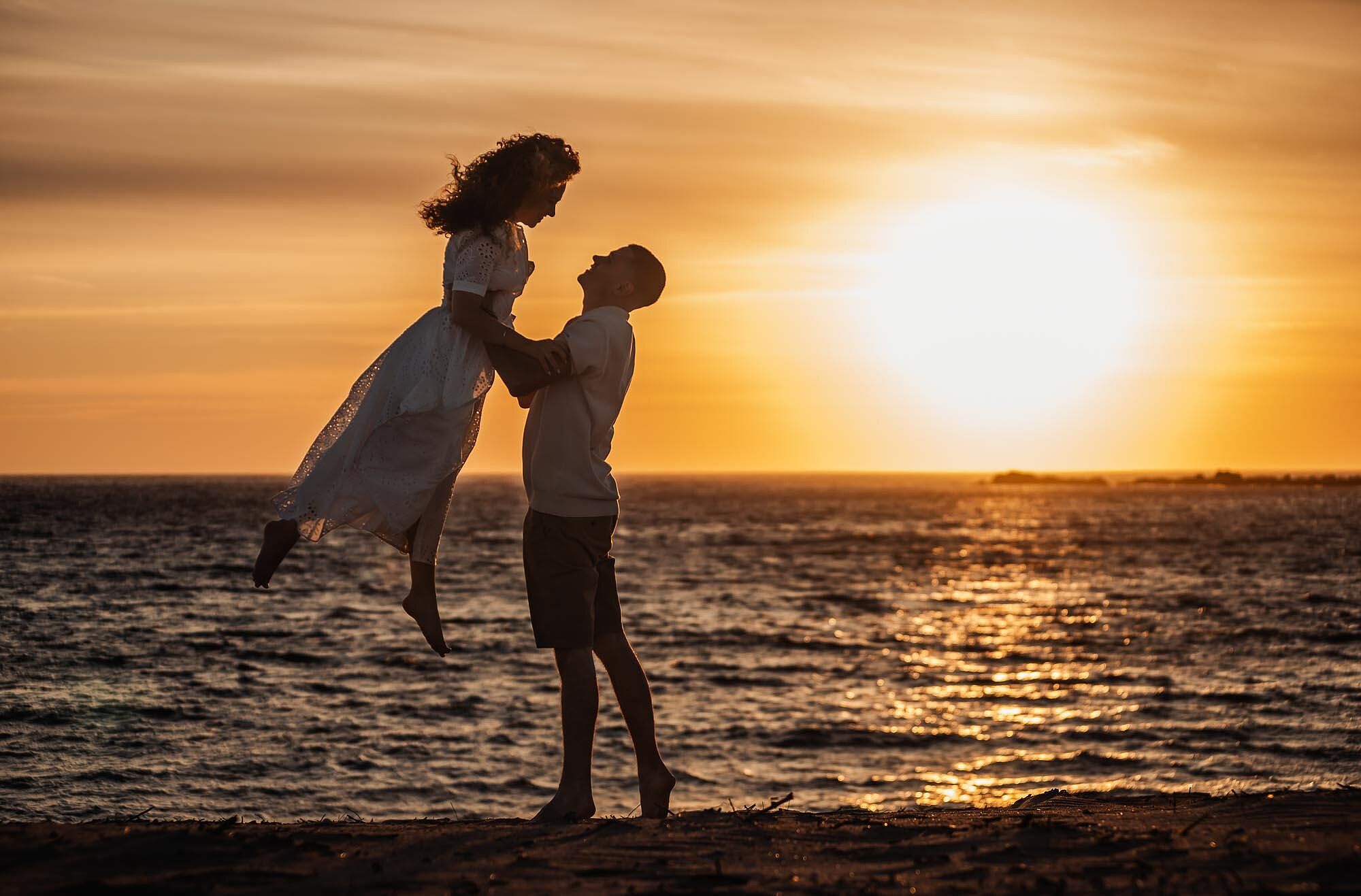 homem levanta a mulher junto ao mar ao pôr do sol