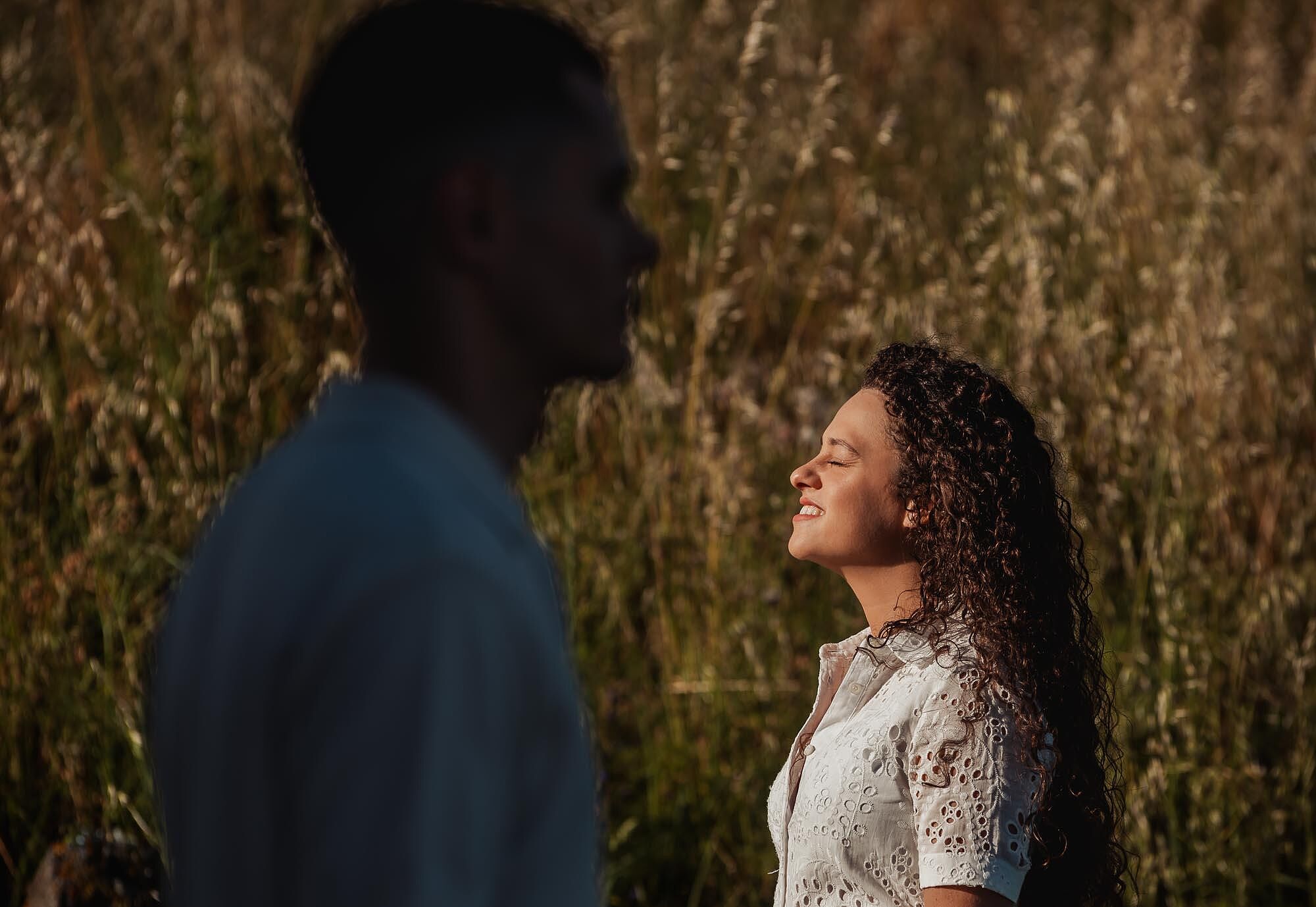 mulher de olhos fechados a sorrir enquanto sente o sol com homem desfocado em primeiro plano