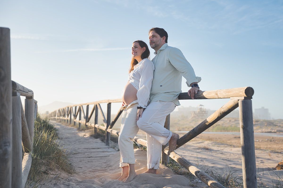 Pareja embarazada apoyada en la valla de madera playa Xeraco - sesión embarazo fotógrafo Safor Juan Reig