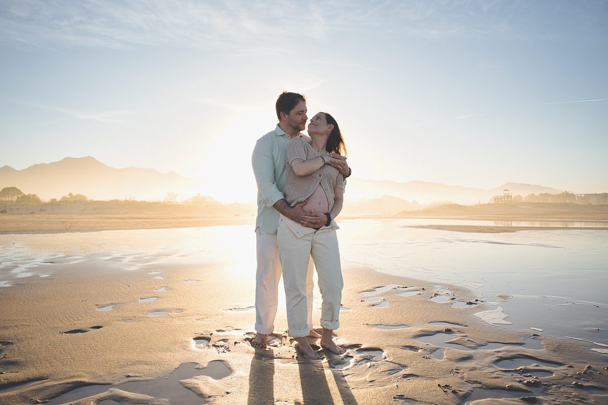 Pareja embarazada abrazada al atardecer en la playa de Xeraco con las montañas al fondo - fotógrafo embarazo Valencia