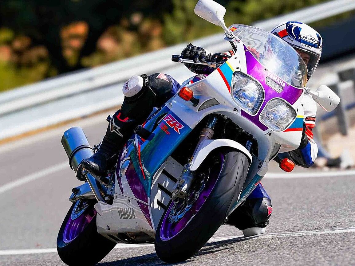 Primer plano de un motociclista con traje y rodilleras blancas, inclinando una motocicleta deportiva clásica (posiblemente una Yamaha FZR) en una curva de carretera. La moto presenta un llamativo diseño retro de colores blanco, púrpura y azul. El piloto lleva el cuerpo descolgado y el neumático delantero está en pleno contacto con el asfalto. Un guardarraíl metálico se ve desenfocado en el fondo.
