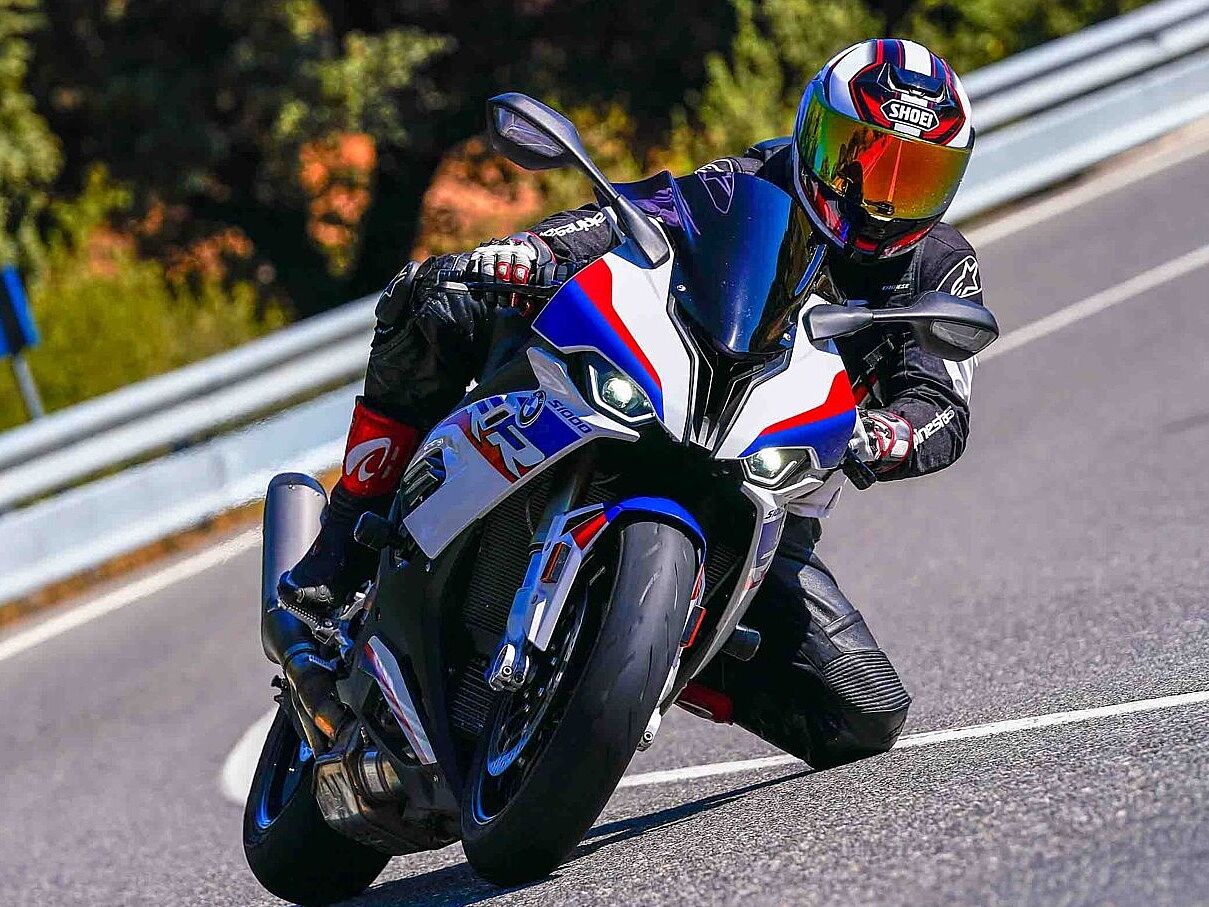 Un motociclista vestido con traje de cuero negro y casco con visera reflectante roja, conduciendo una motocicleta deportiva (posiblemente una BMW S 1000 RR) con diseño en colores blanco, azul y rojo. La moto está muy inclinada en una curva en una carretera asfaltada, y el piloto tiene la rodilla cerca del suelo. El entorno es una carretera soleada con vegetación lateral.
