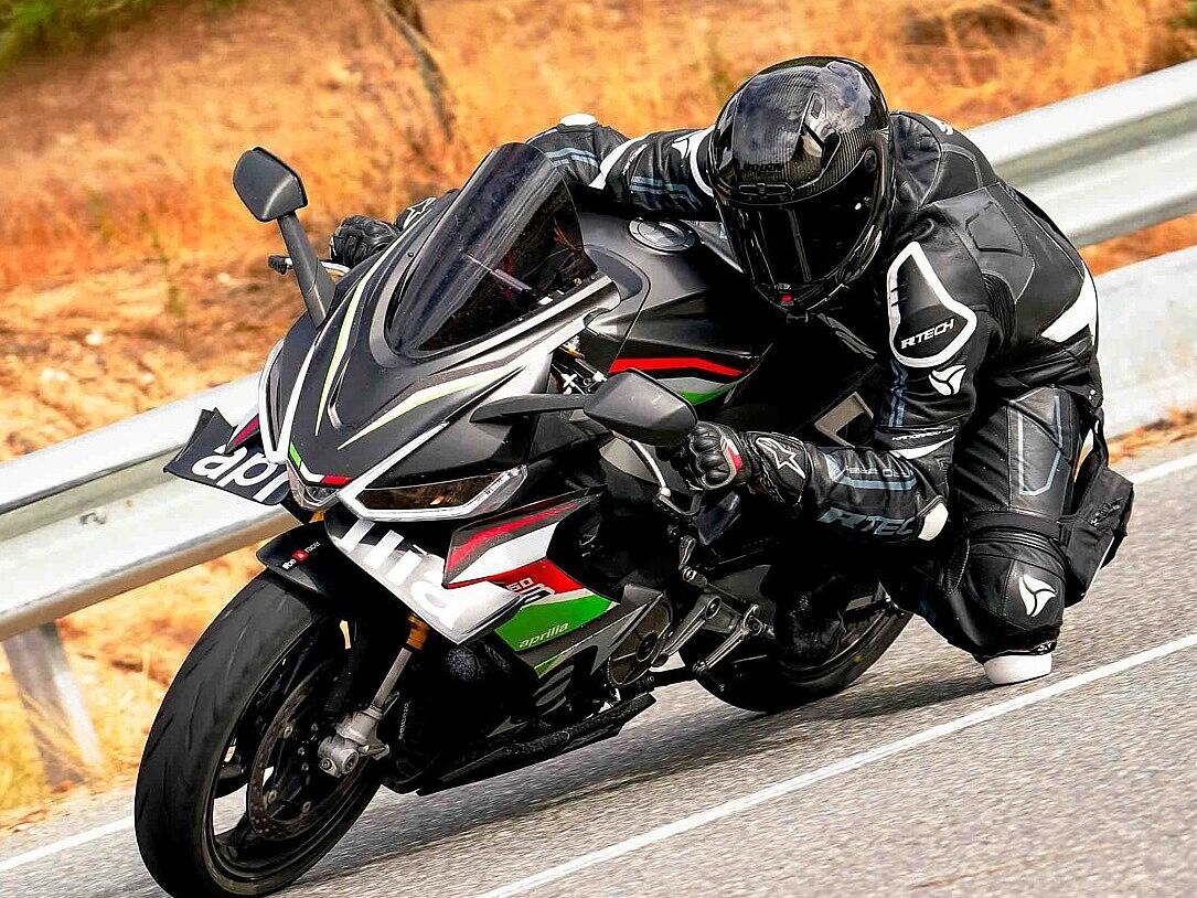 Un motociclista vestido con un traje de cuero negro y casco integral oscuro, conduciendo una motocicleta deportiva negra, blanca, verde y roja (posiblemente una Aprilia), inclinado agresivamente en una curva de carretera. El piloto tiene la rodilla rozando el asfalto. Se aprecia una valla metálica de protección en el lado izquierdo y un entorno natural desenfocado.