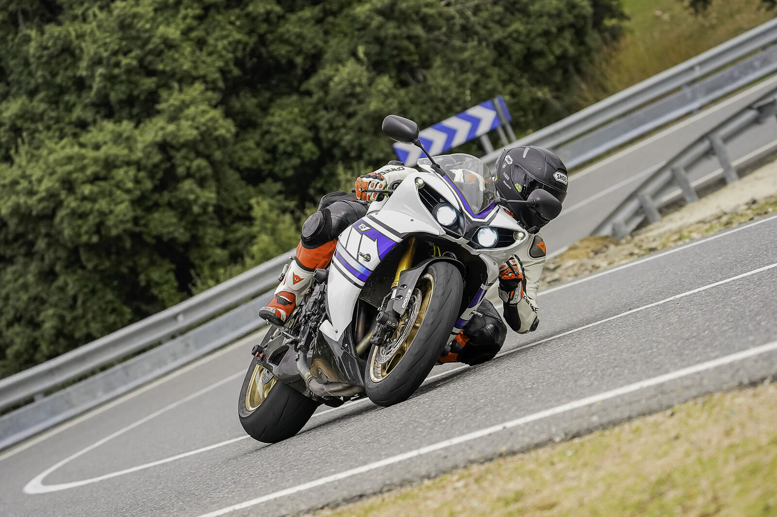 Un piloto con mono Dainese blanco y rojo inclina una Yamaha R1 de colores clásicos en una curva cerrada; se aprecia el contacto de la rodilla con el asfalto y las llantas doradas de la moto.
