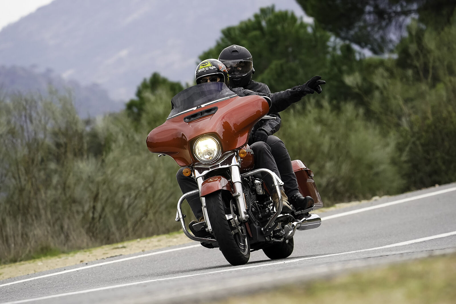 Vista frontal de una motocicleta tipo bagger roja con dos ocupantes en una carretera de montaña; el pasajero señala hacia el horizonte mientras el conductor mantiene la vista al frente.