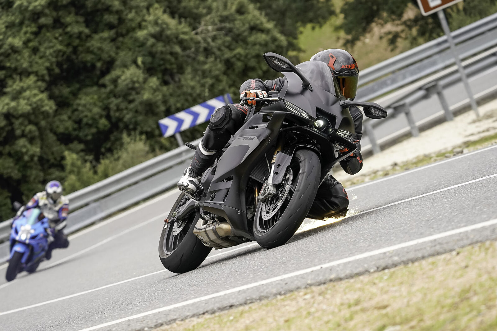 Plano dinámico de un motociclista con mono de competición negro inclinándose agresivamente en una curva de carretera; la rodillera roza el asfalto provocando chispas mientras otra moto azul le sigue de cerca.