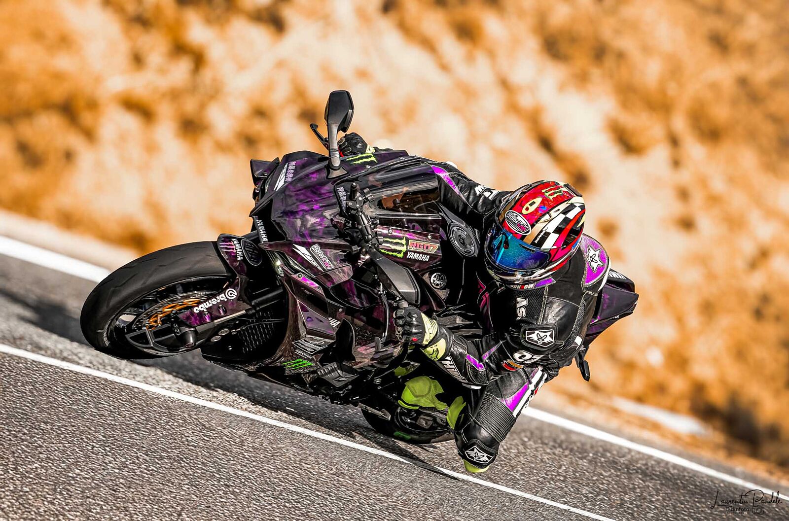 Motocicleta deportiva yamaha r1 m o similar con piloto y pasajero inclinándose en una curva en carretera de montaña. Llevan equipación de moto negra y cascos. El fondo es un talud de vegetación verde y una barrera de seguridad metálica.