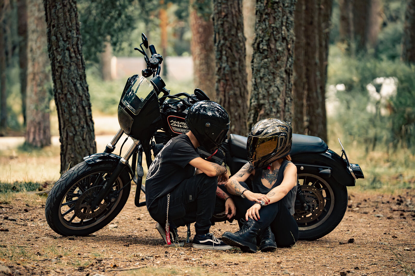Sesion-de-fotos-moto-custom-pareja-madrid Pareja motera con cascos integrales posando junto a una moto Harley-Davidson negra en un bosque, durante una sesión fotográfica de estilo custom y lifestyle con Odinfoto.