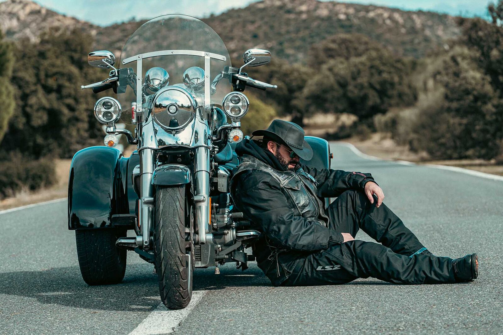 sesion-fotos-trike-harley-davidson-roadster Motorista con sombrero vaquero descansando en el asfalto junto a un trike Harley-Davidson negro de tres ruedas, durante un reportaje de fotografía de estilo custom en carretera con Odinfoto.