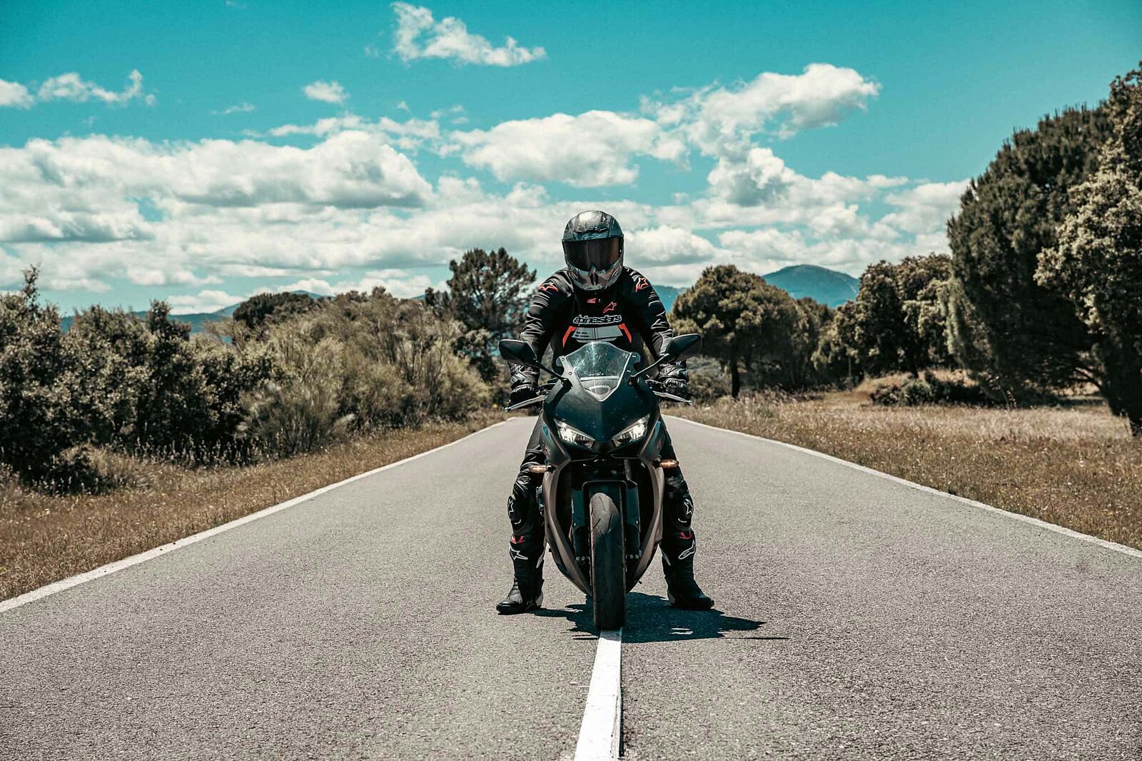 sesion-fotos-moto-deportiva-carretera-madrid Piloto con equipación Alpinestars posando de frente sobre una moto deportiva negra en mitad de una carretera de montaña, durante una sesión fotográfica exclusiva con Odinfoto.