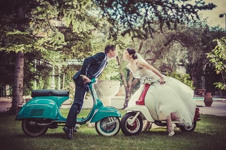 Reportaje Boda Vintage Original | Novios en Scooter Vespa Novio en traje oscuro sobre Vespa turquesa y novia con vestido blanco en Vespa crema inclinándose para besarse en un jardín frondoso.