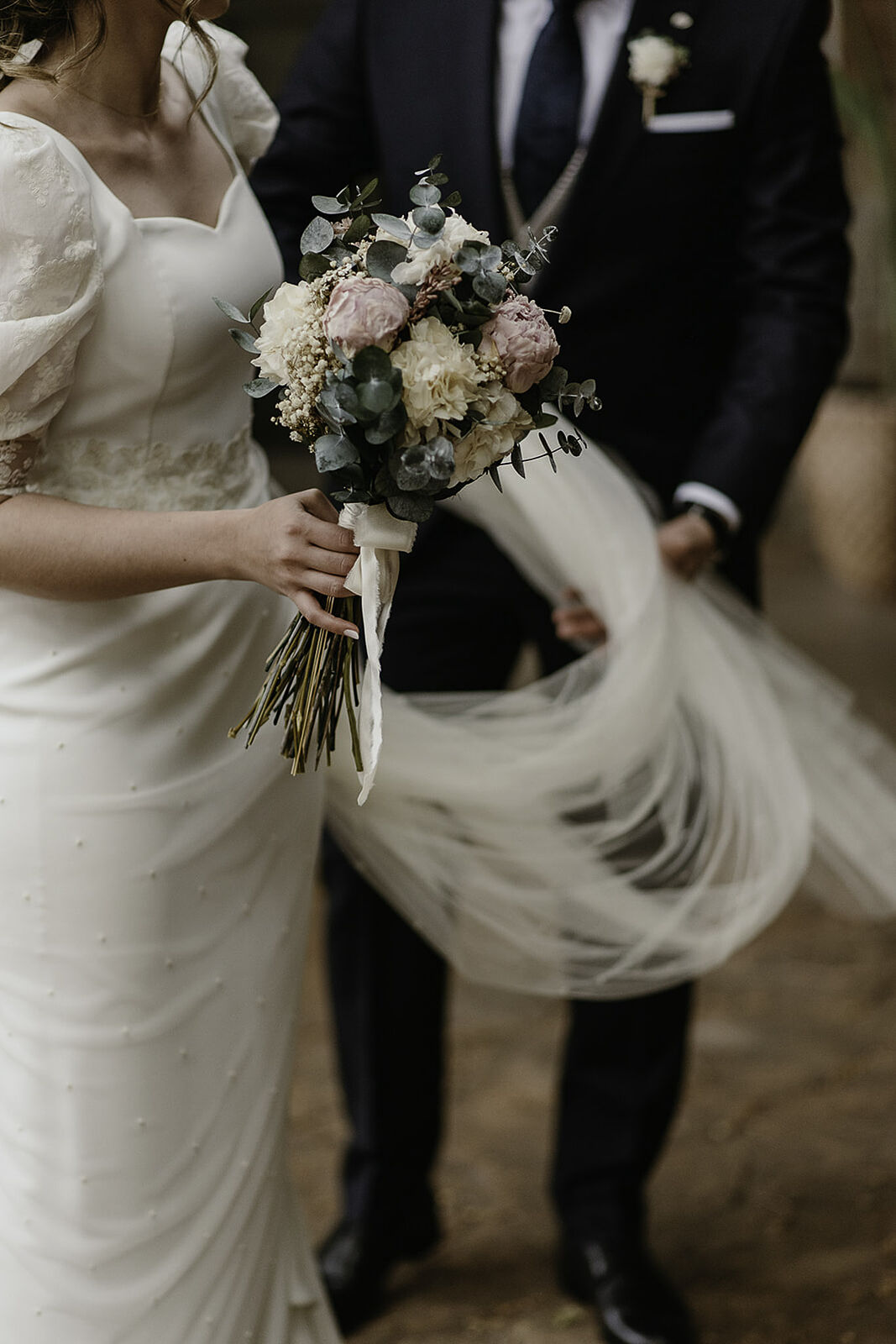 Detalle de las manos de la novia sosteniendo un ramo de peonías y eucalipto, con el velo en movimiento y el novio de fondo. Fotografía profesional en Madrid por Luzdesi.