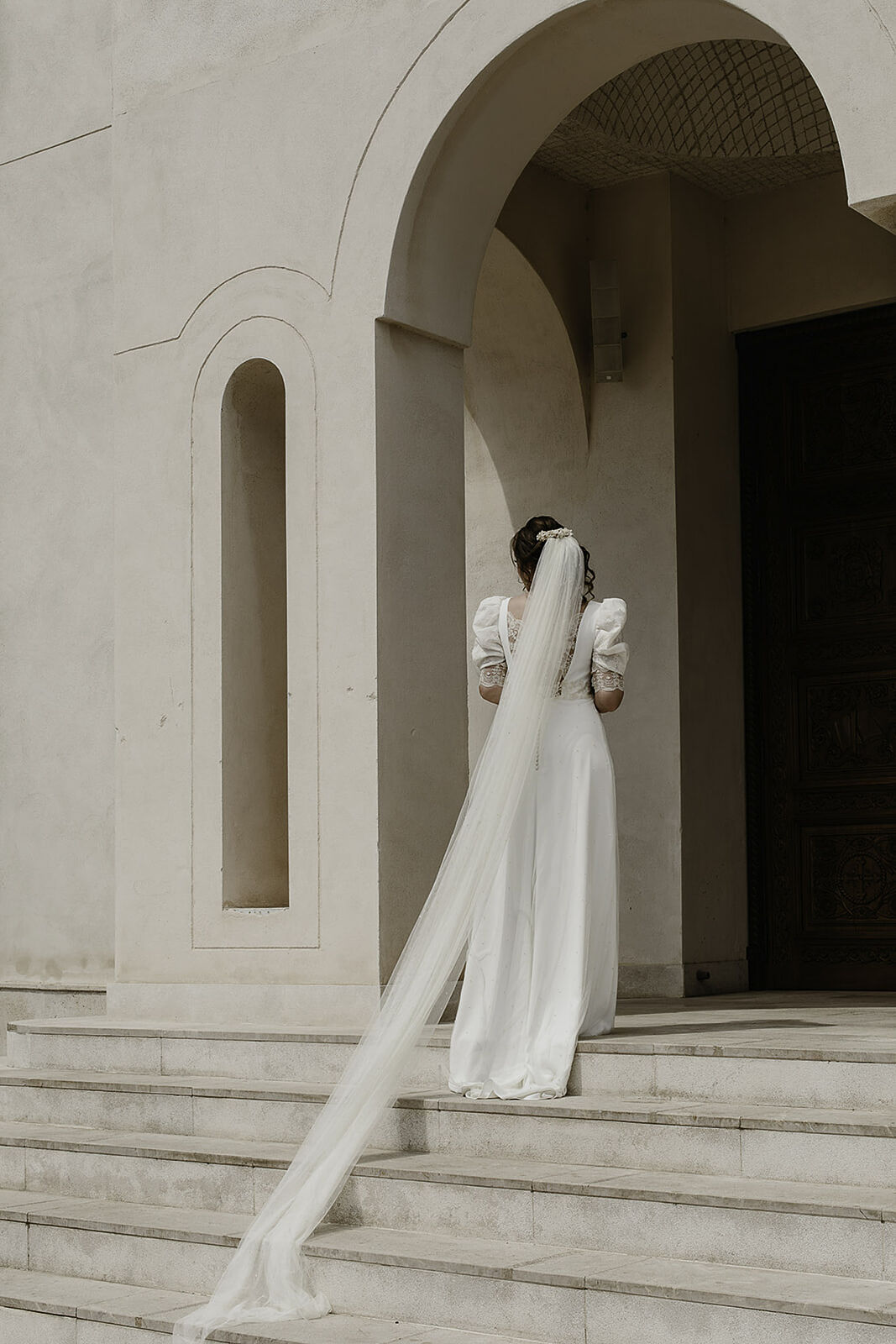 Novia de espaldas subiendo escaleras de piedra hacia un arco, luciendo un velo muy largo y mangas abullonadas. Fotografía de boda elegante en Madrid por Luzdesi.