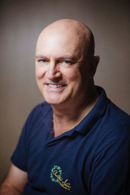 Antonio Siles, fotógrafo profesional en Almería, en un retrato de estudio con camiseta polo azul corporativa, sonriente y con iluminación clásica de estudio sobre fondo marrón neutro.