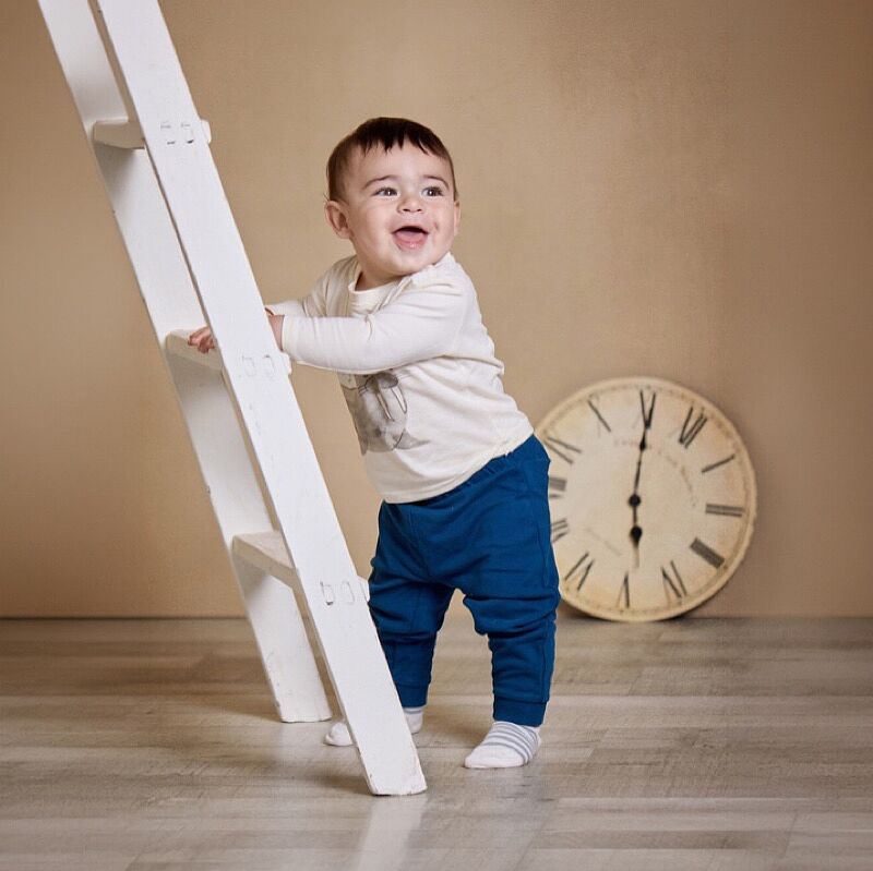Bebé sonriente con camiseta blanca estampada y pantalones azules, apoyado en una escalera de madera blanca en un estudio fotográfico, con un gran reloj de pared vintage al fondo y suelo de madera.