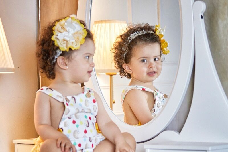 Bebé con diadema amarilla de flores y vestido blanco de lunares de colores, sentada frente a un espejo ovalado blanco, mirándose y reflejándose en él, con una lámpara de mesa encendida al fondo en un estudio fotográfico.