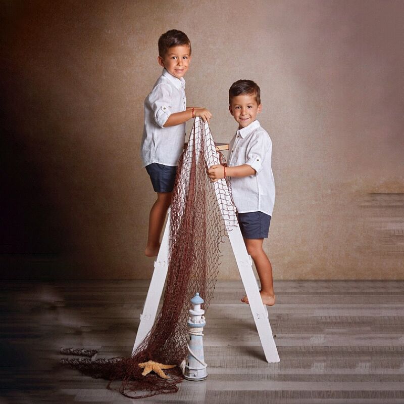Dos niños sonrientes con camisas blancas de lino y shorts azules, posando descalzos sobre una escalera de madera blanca con una red de pesca, un faro decorativo y una estrella de mar, en un estudio fotográfico con fondo marrón.
