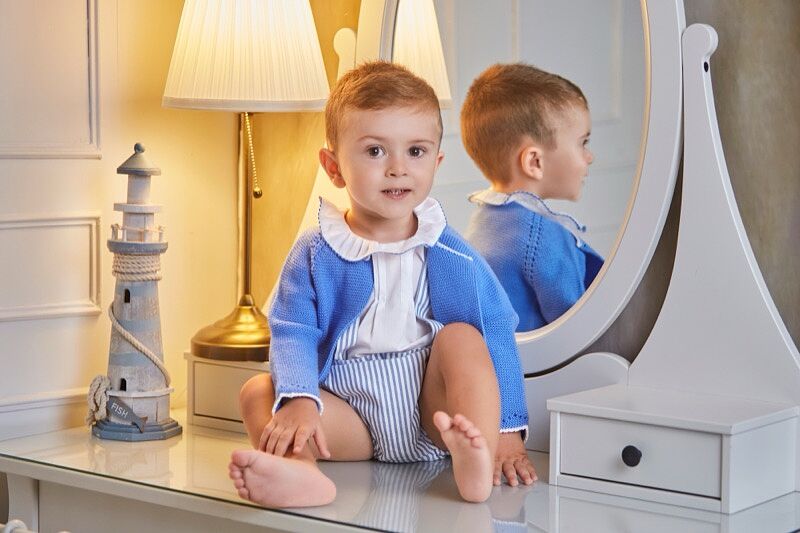 Bebé sonriente con conjunto de punto azul y blanco con cuello de volantes, sentado descalzo sobre la superficie de cristal de un tocador blanco, con un espejo ovalado donde se refleja, un faro decorativo y una lámpara de mesa encendida al fondo.