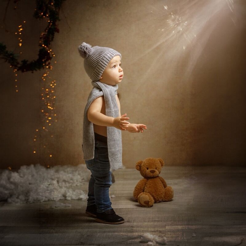 Niño pequeño con gorro de lana gris con pompón, bufanda a juego y jeans, de pie en un estudio fotográfico con suelo de madera y nieve artificial, observando un haz de luz mientras un osito de peluche marrón descansa en el suelo junto a él, con luces navideñas al fondo.